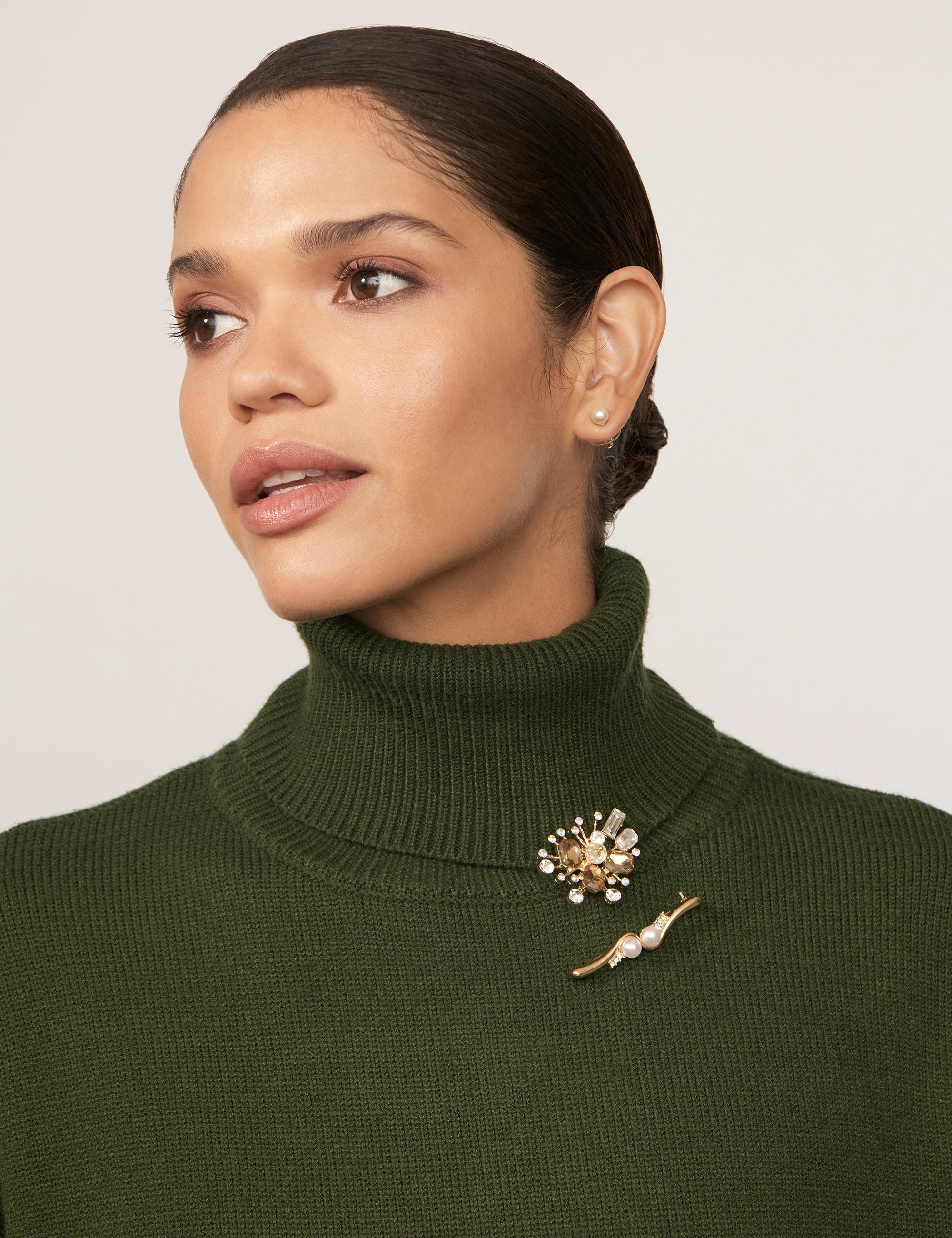 A woman with tan skin and dark hair wears a green turtleneck sweater. She is wearing a pair of sparkling brooches with gold-coloured plating.