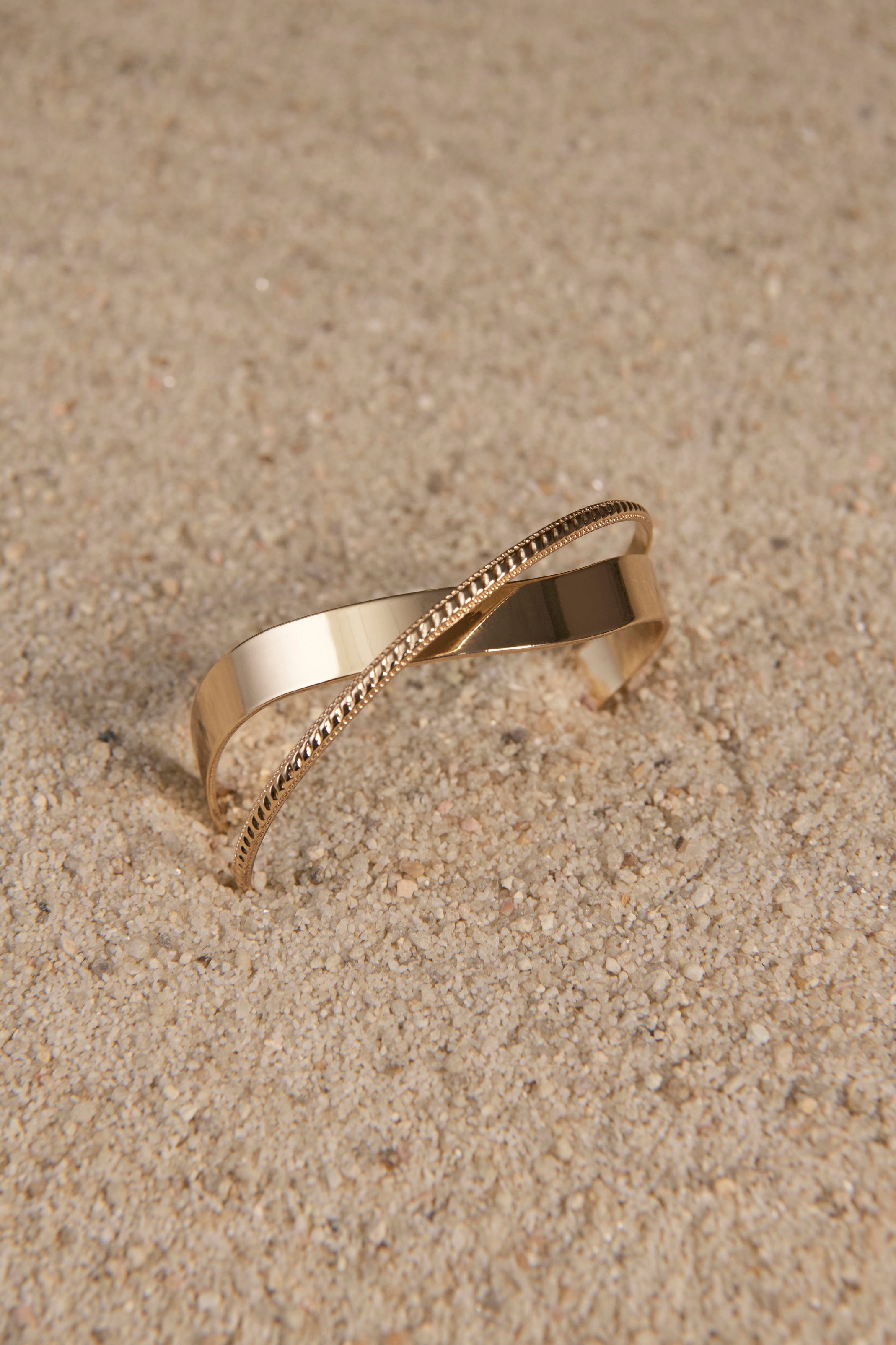 A gold bangle with crossed dual bands sits on a sandy background