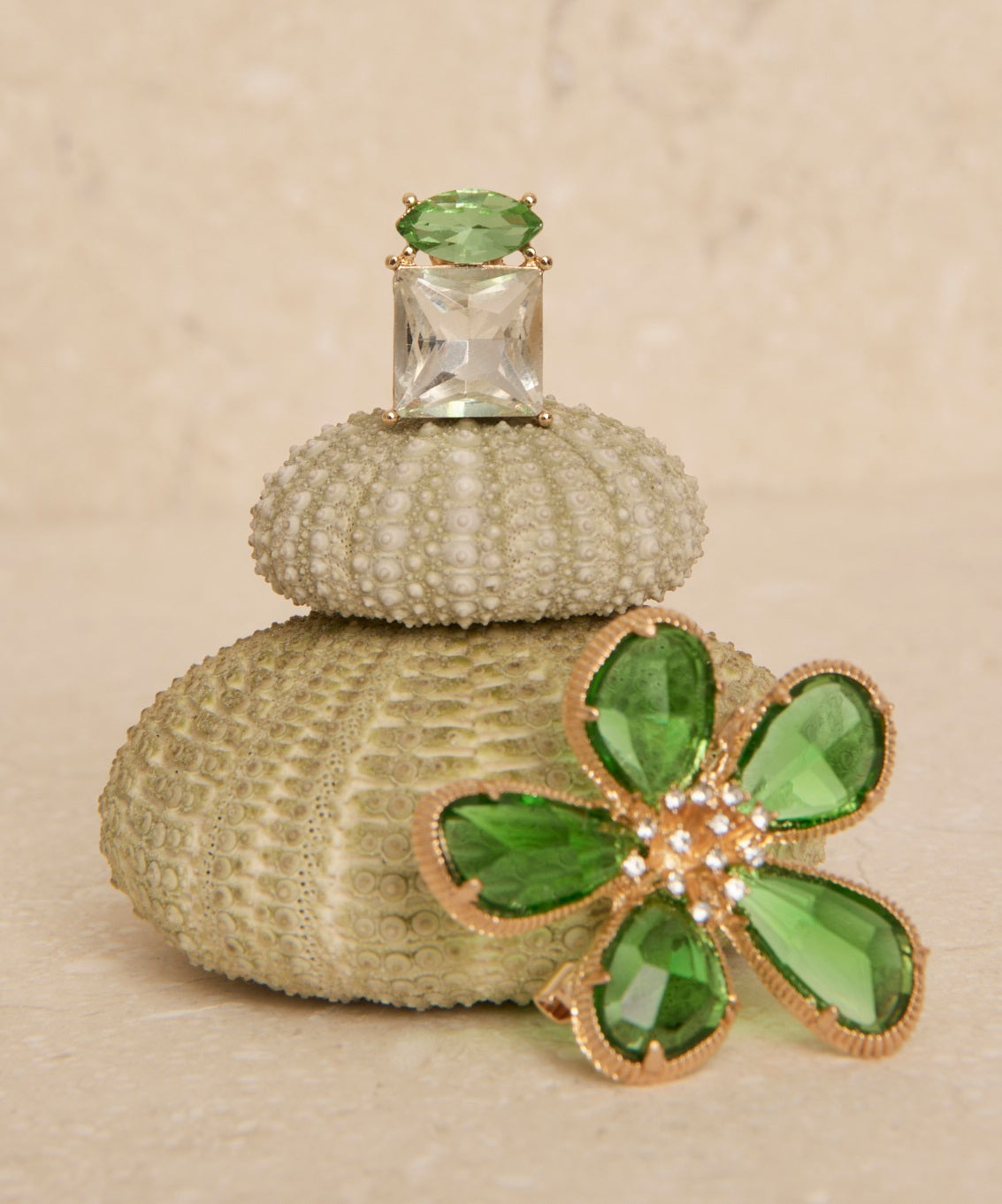 A flower-shaped brooch with gold-coloured plating and faceted green stone petals rests against a rock. On top of the rock is a single earring with a clear faceted square stone and a green oval stone accent. 