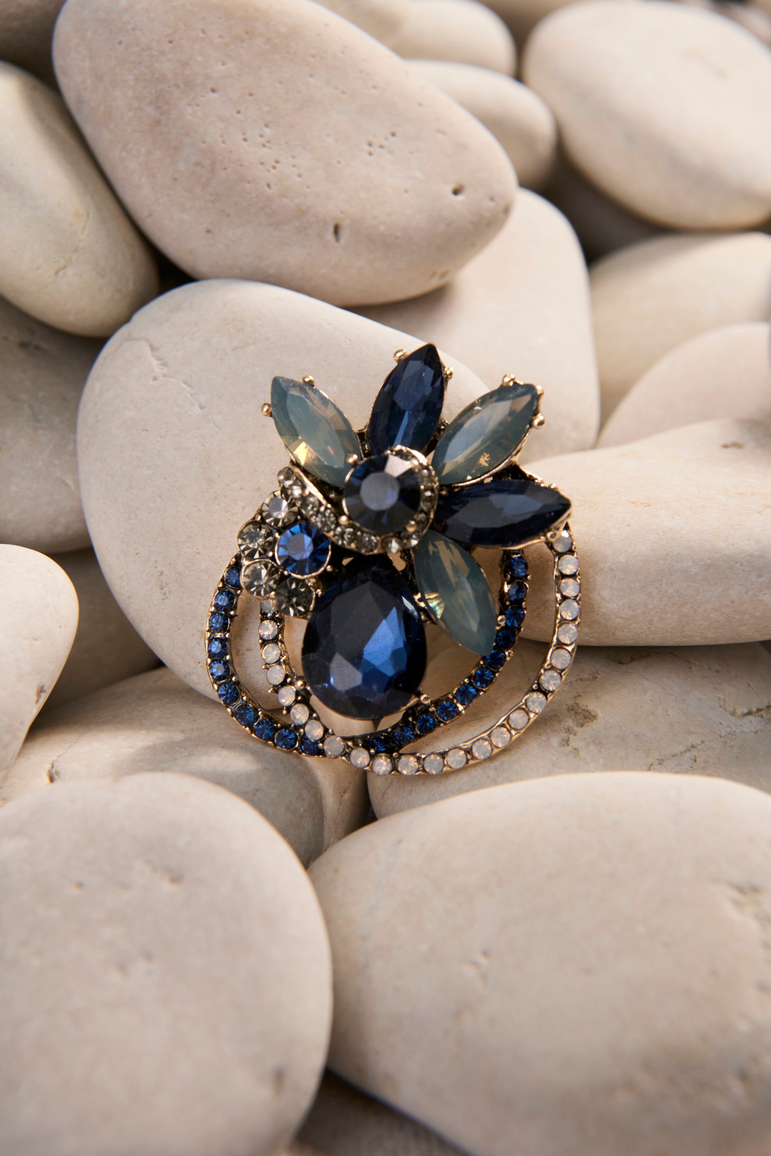 The Coil Petal Brooch sits on a bed of smooth white tones. The brooch features an array of faceted blue stones with fine sparkling stone detailing. 